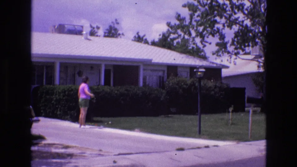 1973 New Mexico home-movie frame showing an adult woman near a concrete driveway, hedges, lawn, lamppost, and single-story suburban house.