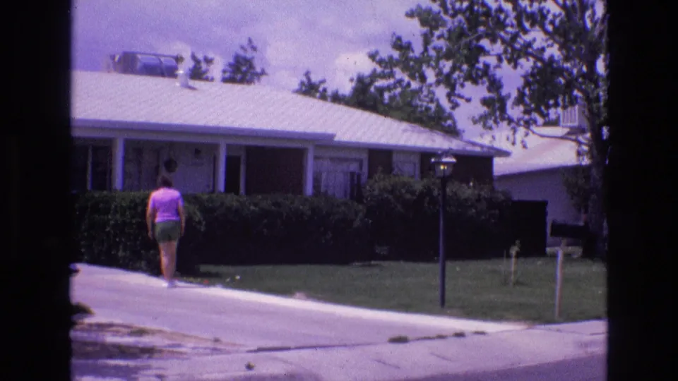 1973 New Mexico home-movie frame showing the woman from behind as she walks along a residential driveway bordered by lawn, hedges, and a single-story house.