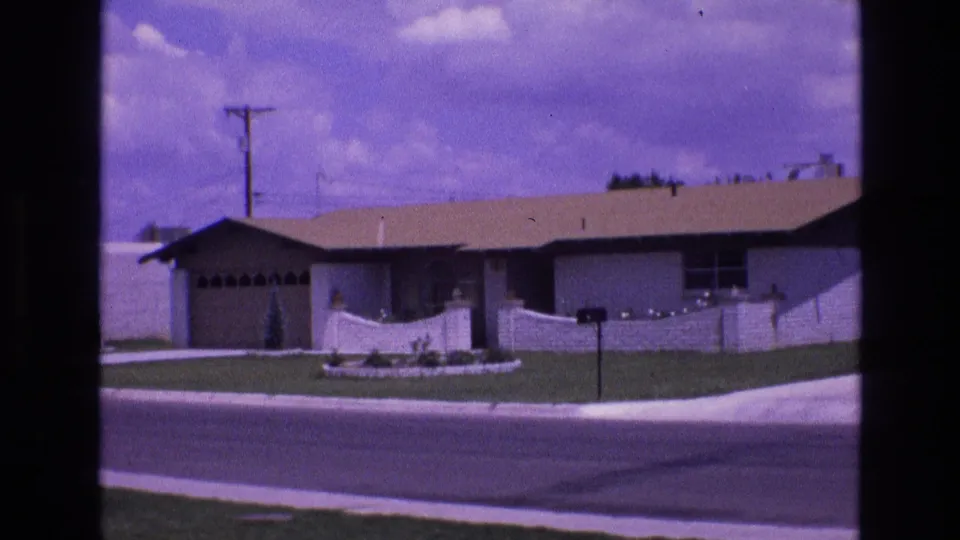 1973 New Mexico home-movie frame showing a single-story suburban house across the street with a garage, front lawn, mailbox, and low white fence.