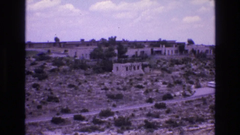 1973 New Mexico 8mm frame showing undeveloped desert scrub, a curving road, low distant buildings, and open sky.