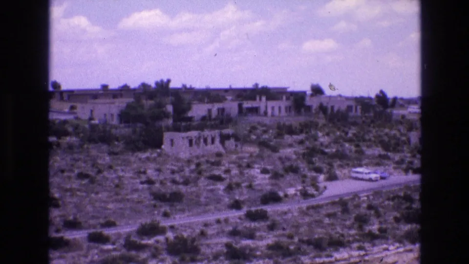 1973 New Mexico 8mm frame showing scrub-covered terrain, a road, distant low structures, and a broad partly cloudy sky.