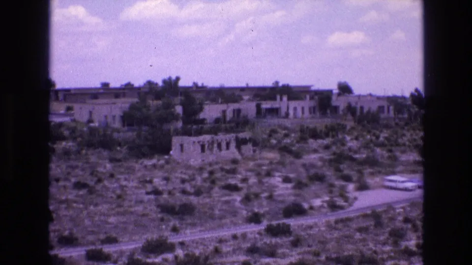 1973 New Mexico 8mm frame showing a weathered stone ruin with window openings on a scrub hillside below long low adobe-style buildings.