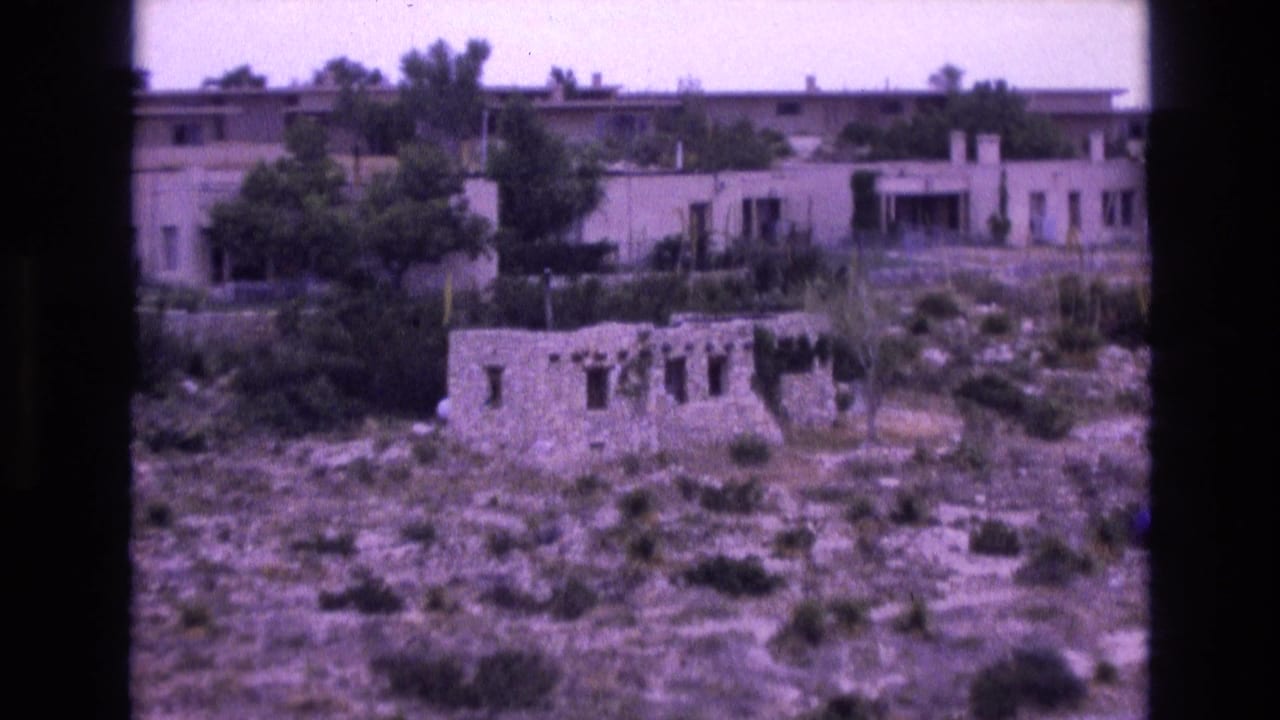 Stone ruin and low buildings seen across a dry, brush-covered slope.