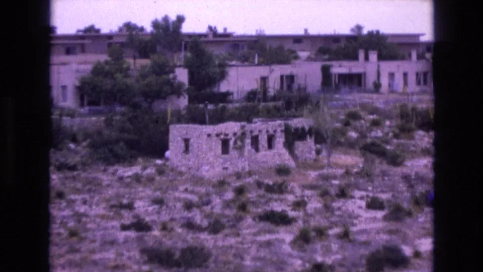 1973 New Mexico 8mm frame with a closer view of a weathered stone ruin in front of low adobe-style buildings and trees on a dry hillside.