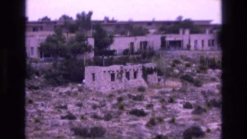 1973 New Mexico 8mm frame showing low adobe-style buildings behind a weathered stone ruin, with desert brush and trees across the slope.