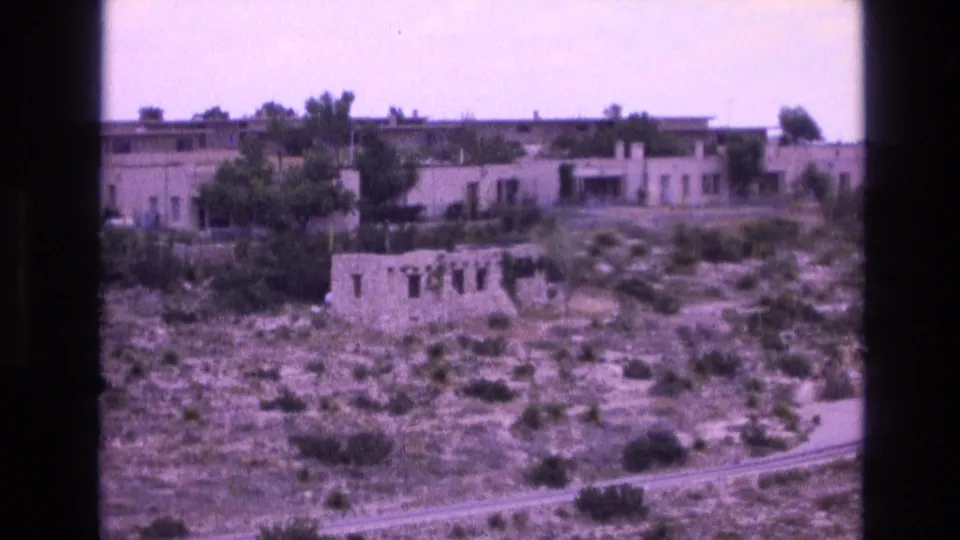1973 New Mexico 8mm frame showing a weathered stone ruin on a dry hillside, low adobe-style buildings behind it, and a curving road near the lower edge.