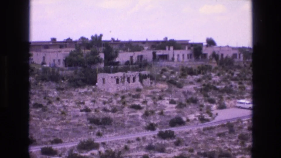 1973 New Mexico home-movie frame showing low stucco-style structures across dry scrub terrain with a road crossing the foreground.