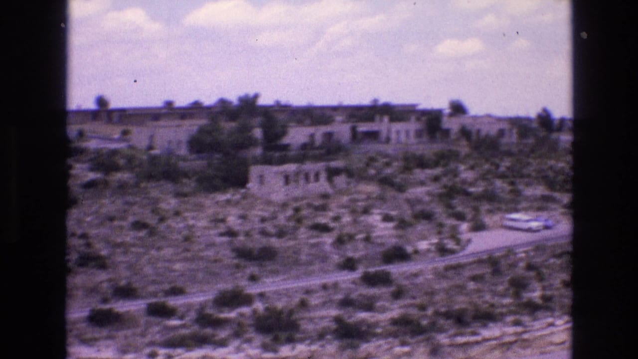 Distant desert buildings and scrubland viewed across a road beneath scattered clouds.