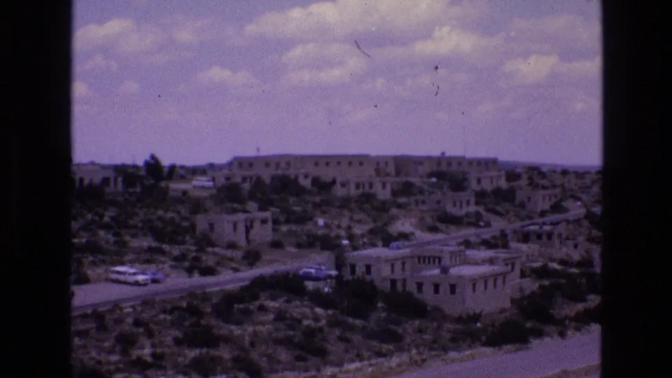 Wide 1973 New Mexico home-movie view of stucco-style buildings, a curving road, and vehicles moving through a dry settlement landscape.