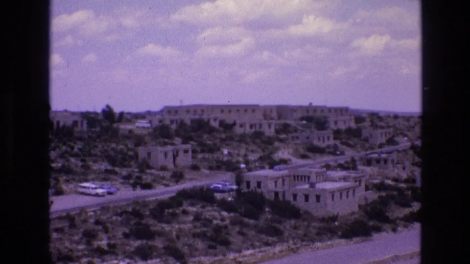 1973 New Mexico frame showing a dry settlement with low stucco-style structures, a road, and visible vehicles under scattered clouds.
