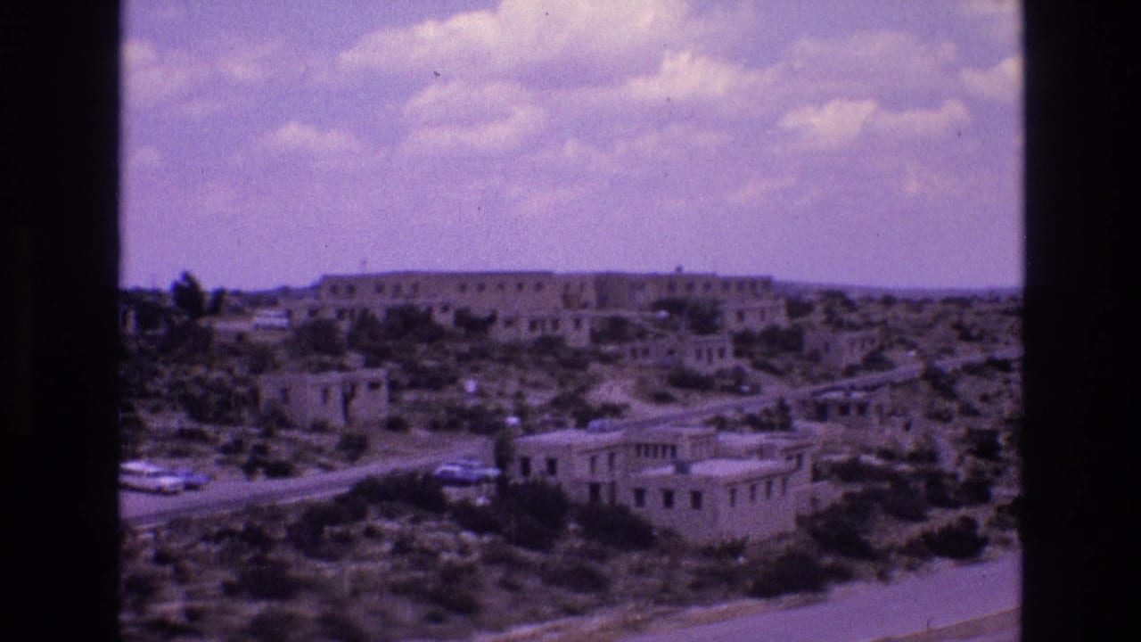Distant desert village with low buildings, shrubs, road, cars, and cloudy sky.