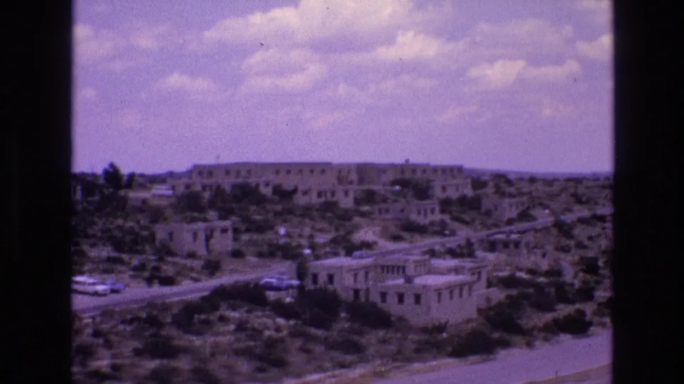 Wide 1973 8mm view of a New Mexico desert settlement with low adobe-like buildings, sparse shrubs, a road, small vehicles, and a clouded sky.