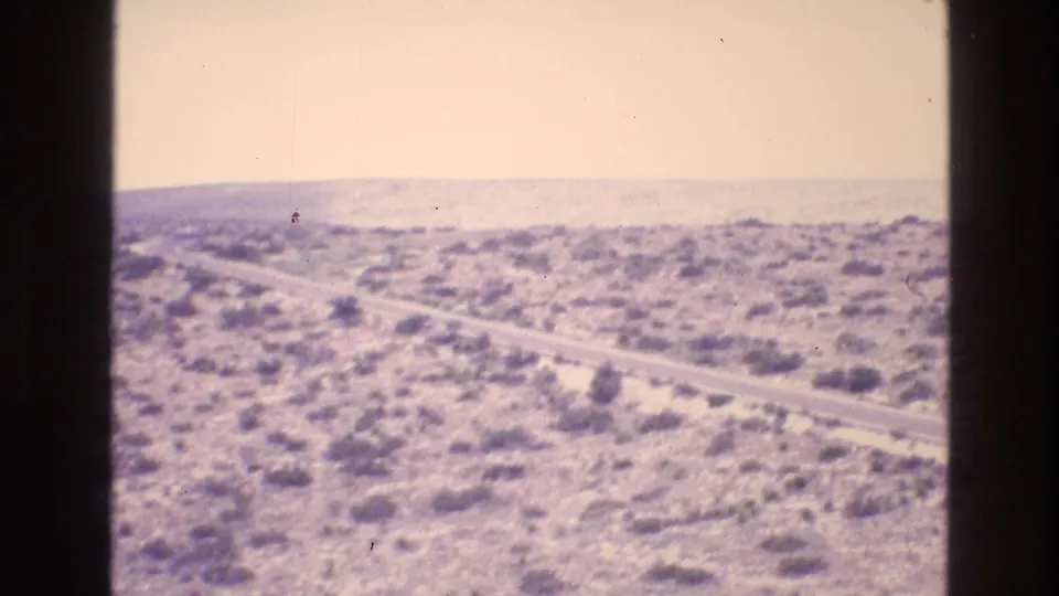 Overexposed 8mm frame of New Mexico desert scrubland with a road running diagonally toward a hazy horizon.