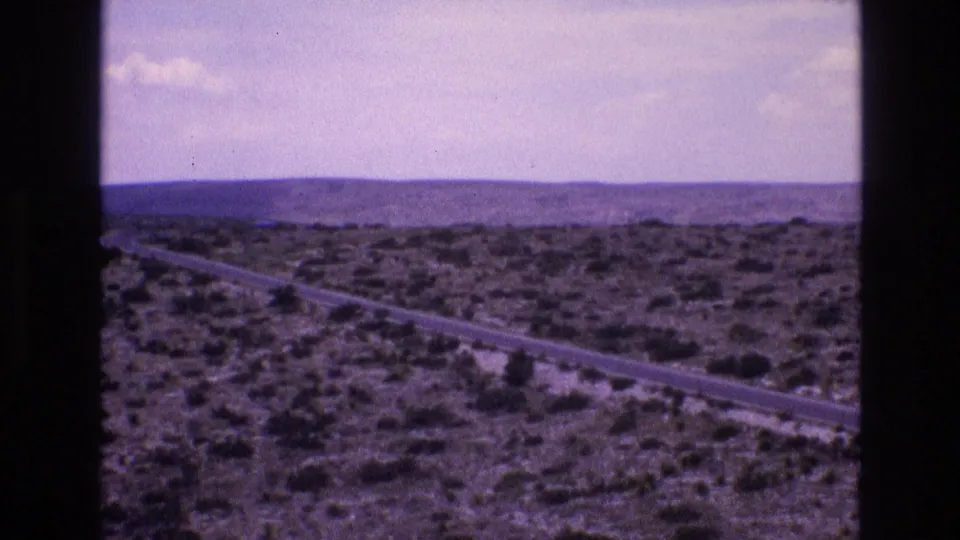 Wide 1973 8mm New Mexico landscape showing a road through dark desert scrub, distant low hills, and an open evening sky.
