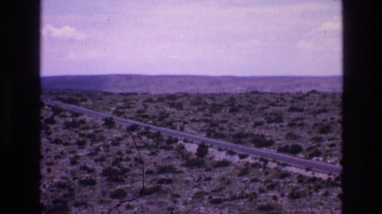 1973 New Mexico 8mm frame showing a paved road through arid scrubland with low hills on the horizon and faded home-movie color.