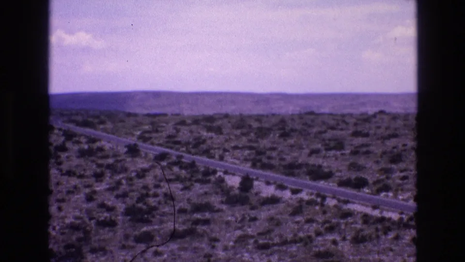 1973 New Mexico 8mm frame showing a paved road through arid scrubland with low hills on the horizon and faded home-movie color.