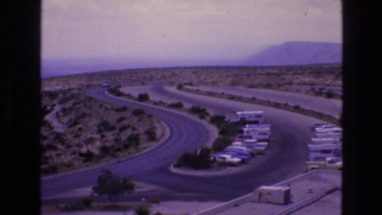 1973 New Mexico travel frame showing a curving access road, parked vehicles and RVs, desert vegetation, and low hills in the distance.