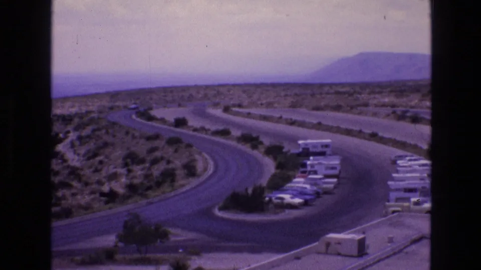 1973 New Mexico travel frame showing a curving access road, parked vehicles and RVs, desert vegetation, and low hills in the distance.