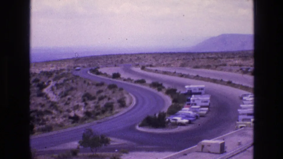 1973 New Mexico 8mm frame with a winding road, roadside parking, cars and RVs, scrub-covered terrain, and a distant hill.