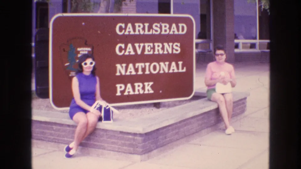 1973 New Mexico home-movie frame showing two women in 1970s attire seated beside a brown Carlsbad Caverns National Park sign near a building entrance.