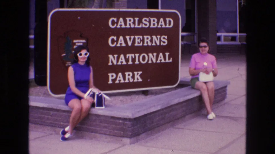 Two adult women in 1970s-style summer clothing sit on a stone bench beside the Carlsbad Caverns National Park sign in New Mexico.