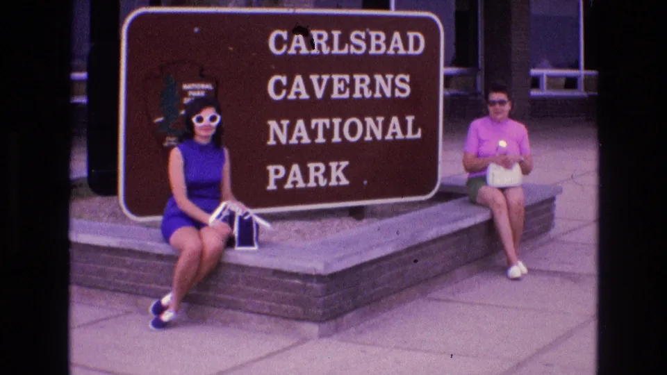 Two adult women sit for a 1973 home-movie shot near the Carlsbad Caverns National Park sign, with a visitor-area building behind them.