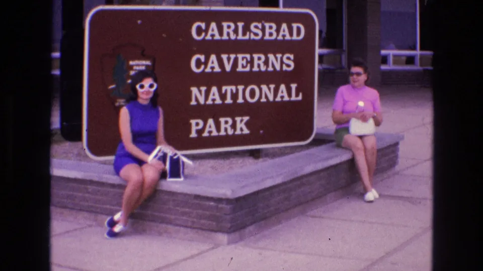 1973 home-movie frame of two women in 1970s clothing seated beside the Carlsbad Caverns National Park sign in New Mexico.
