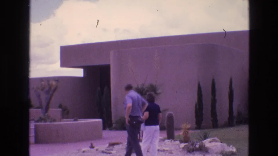 1973 New Mexico park visitor scene with two adults walking toward a modernist adobe-style building bordered by cacti, rocks, and desert landscaping.