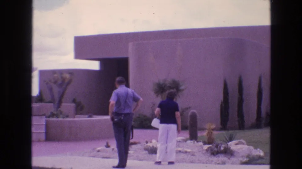 Home-movie frame of two adults walking away from the camera near a low public building and desert landscaping at Carlsbad Caverns National Park.