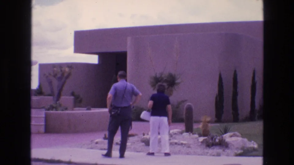 In 1973 New Mexico 8mm footage, a man and woman are seen from behind walking toward a modernist adobe-style building with desert plants and low walls.