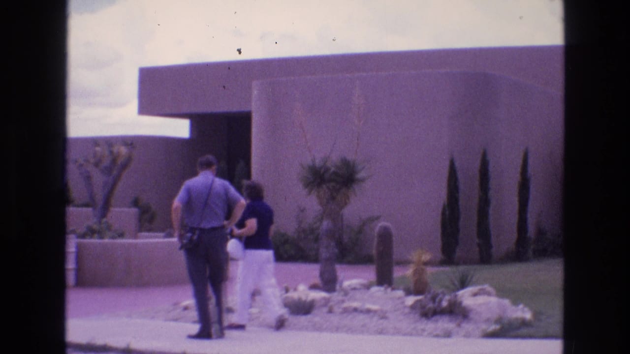 A man and woman continue along the path beside desert landscaping and a modernist building.