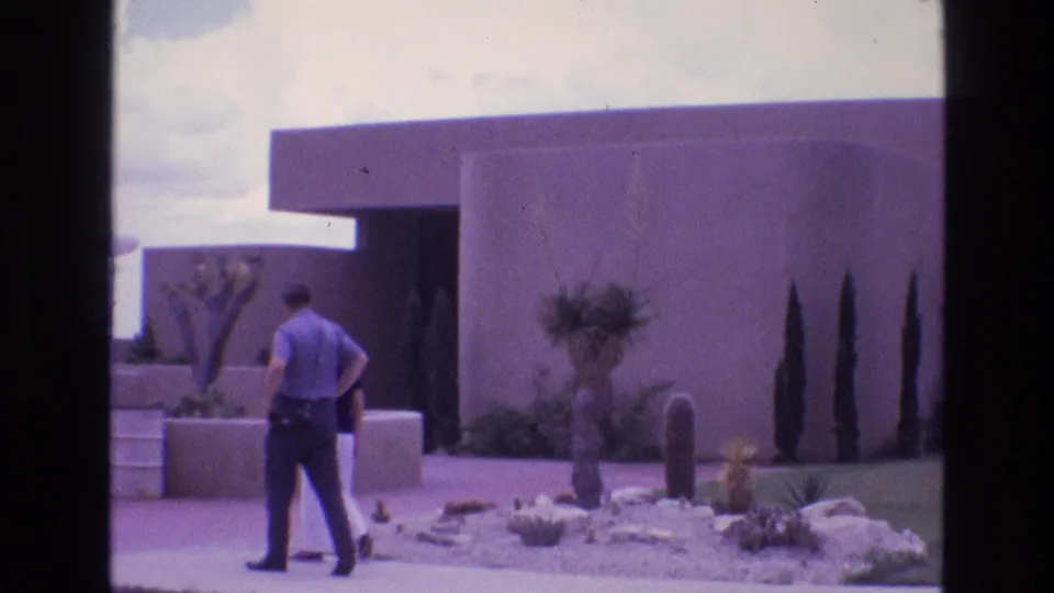 In the 1973 home-movie frame, the man pauses or turns near the walkway while the woman is partly obscured by the low wall of the adobe-style property.