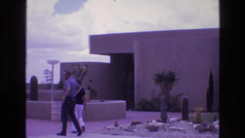The final frame shows the man and woman moving across the foreground of a New Mexico desert garden, with modernist adobe-style architecture and a water tower-like structure in the distance.