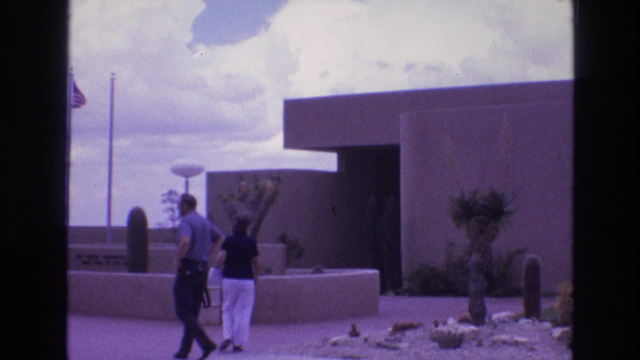 Two adult visitors walk toward the New Mexico Zoological-Botanical State Park entrance area in 1973 home-movie footage, with a low modernist building, desert plants, flagpoles, and cloudy sky visible.