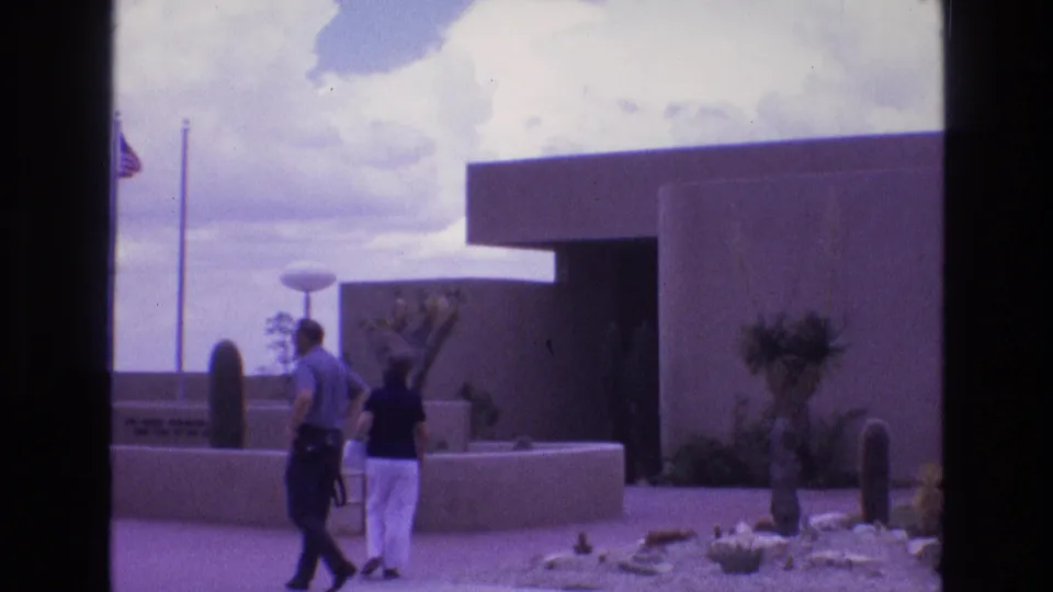 Two adult visitors walk toward the New Mexico Zoological-Botanical State Park entrance area in 1973 home-movie footage, with a low modernist building, desert plants, flagpoles, and cloudy sky visible.