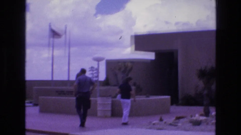 Two visitors continue toward the entrance of the New Mexico zoological-botanical park, passing low stucco walls, planted desert landscaping, flagpoles, and a mid-century modern exterior.