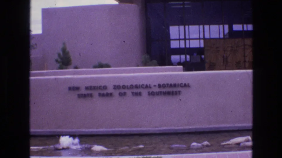 The readable entrance sign for the New Mexico Zoological-Botanical State Park of the Southwest appears in front of a modernist building, with a small water feature and desert landscaping in the foreground.