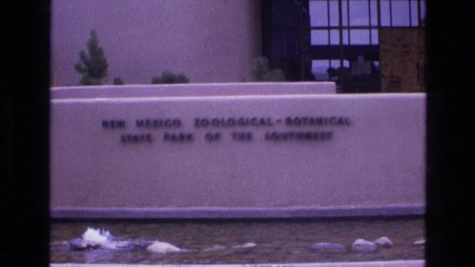 The New Mexico Zoological-Botanical State Park of the Southwest sign fills the lower frame while fountain water, rocks, desert plants, and the entrance building remain visible behind it.