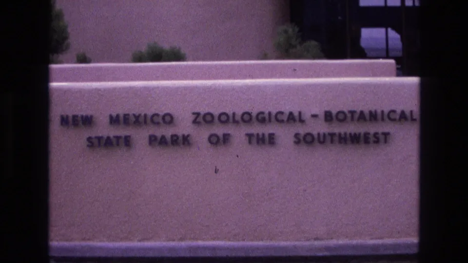 1973 New Mexico home-movie frame showing the engraved New Mexico Zoological-Botanical State Park of the Southwest sign across a pale entrance wall.