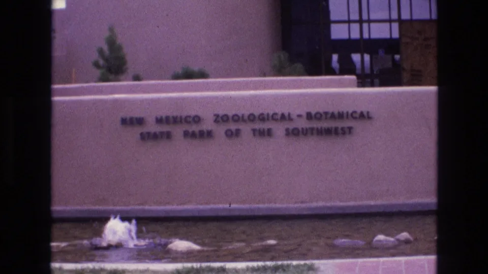 1973 New Mexico zoo-botanical park sign viewed across a shallow fountain area, with stucco architecture and plants behind the wall.