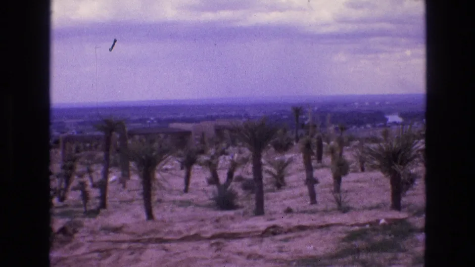 1973 New Mexico 8mm frame with a wide elevated view over sandy terrain, scattered desert plants, faint distant structures, and a flat horizon beneath soft clouds.