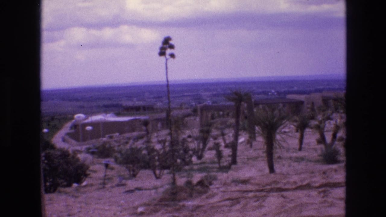 1973 New Mexico 8mm frame showing a dry desert slope with sparse vegetation, a curving road or path, distant structures, and hazy sky.