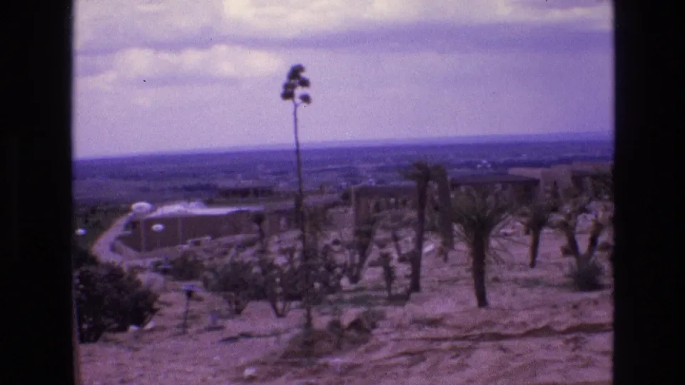 1973 New Mexico 8mm frame showing a dry desert slope with sparse vegetation, a curving road or path, distant structures, and hazy sky.