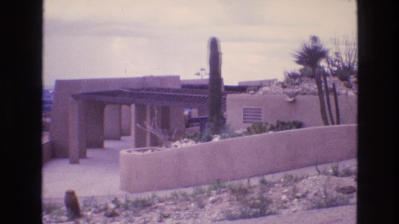 A Southwestern adobe-style house exterior appears beyond a low wall, with cactus and desert landscaping under an overcast sky in 1973 New Mexico footage.