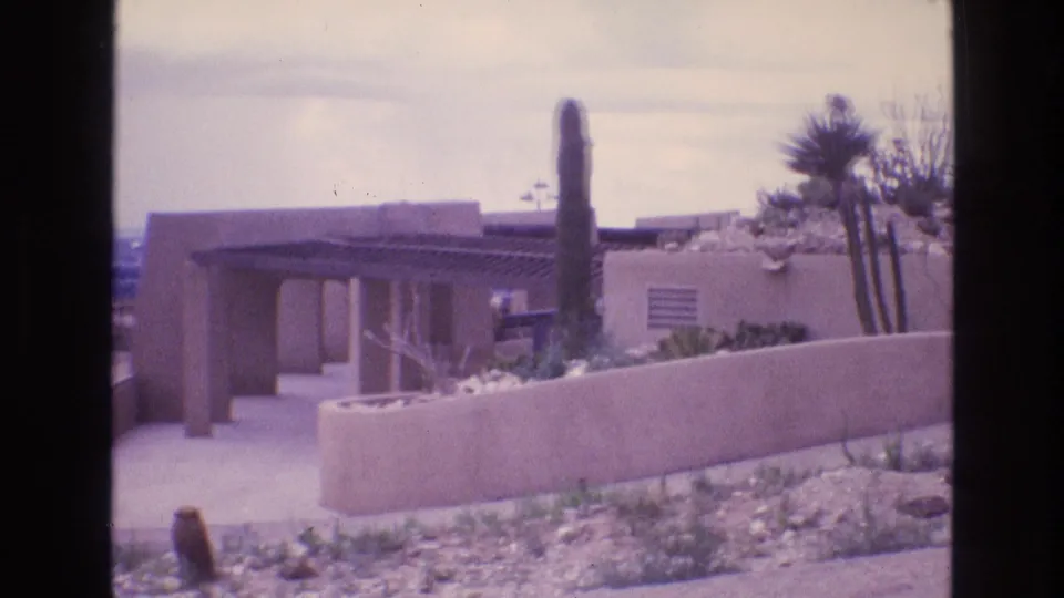A Southwestern adobe-style house exterior appears beyond a low wall, with cactus and desert landscaping under an overcast sky in 1973 New Mexico footage.