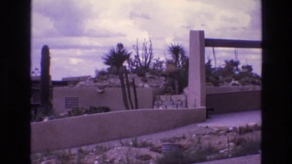 A wider home-movie view shows an adobe-style exterior, cactus plantings, stone landscaping, and a shaded structural element at the edge of the frame in 1973 New Mexico.