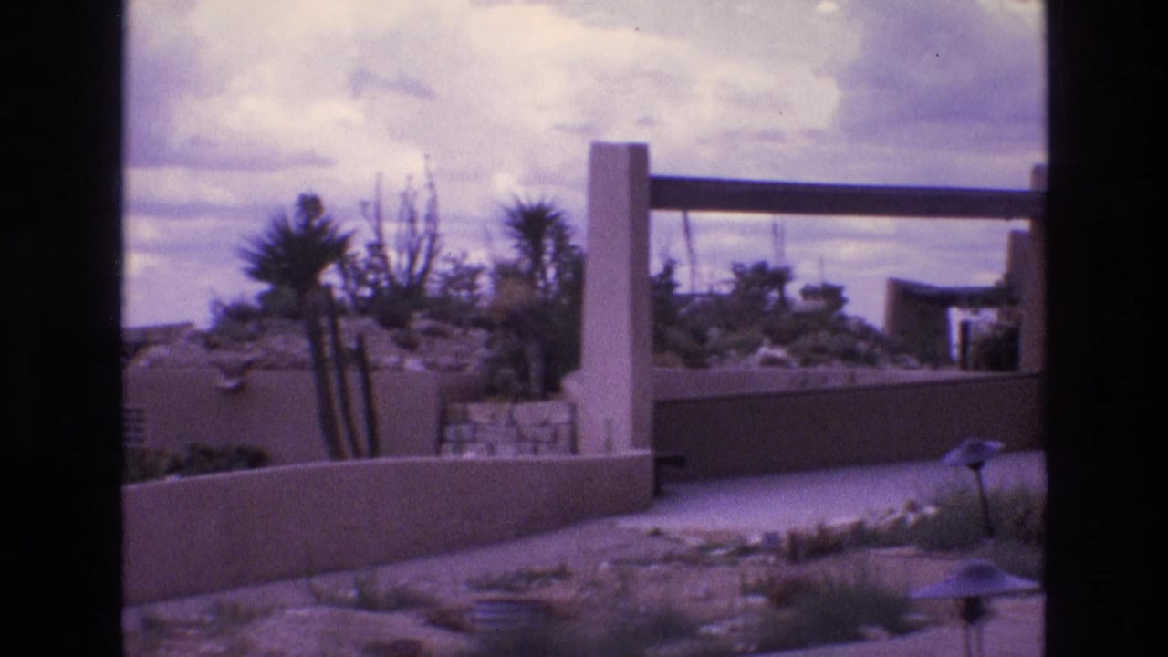 Adobe-style desert house exterior with low walls, yucca-like plants, and cloudy sky.