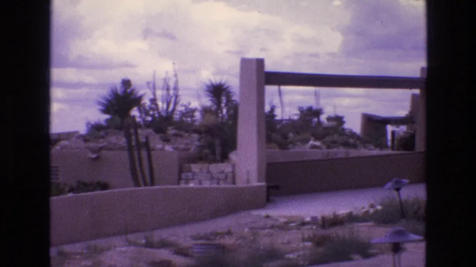 1973 New Mexico 8mm frame showing an adobe-style stucco house exterior behind low walls, with sparse desert planting and a bright clouded sky.