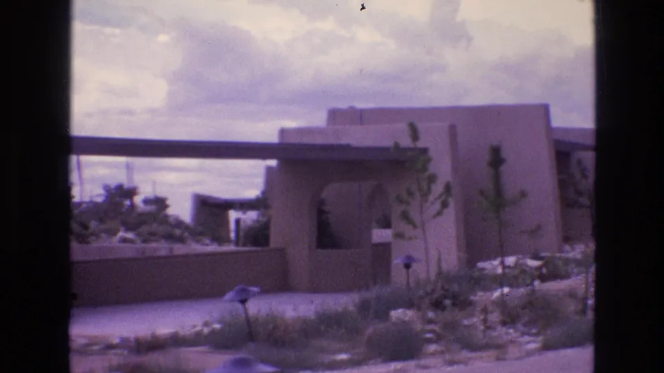 1973 New Mexico 8mm frame showing a flat-roof stucco house, low retaining wall, sparse desert yard, and small path light in soft faded color.