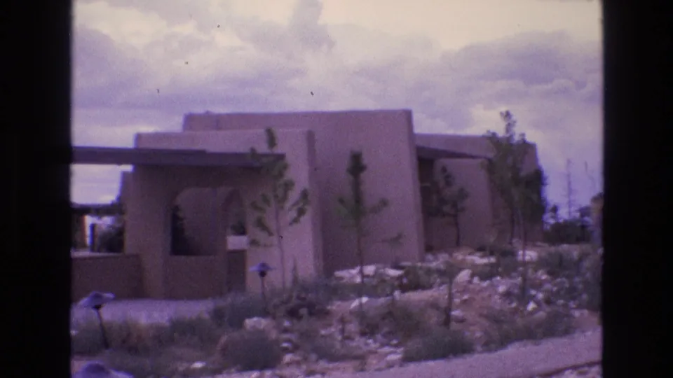 1973 New Mexico home-movie frame of an adobe-style stucco house exterior, rocky desert landscaping, low walls, and sparse plants beneath a bright cloudy sky.